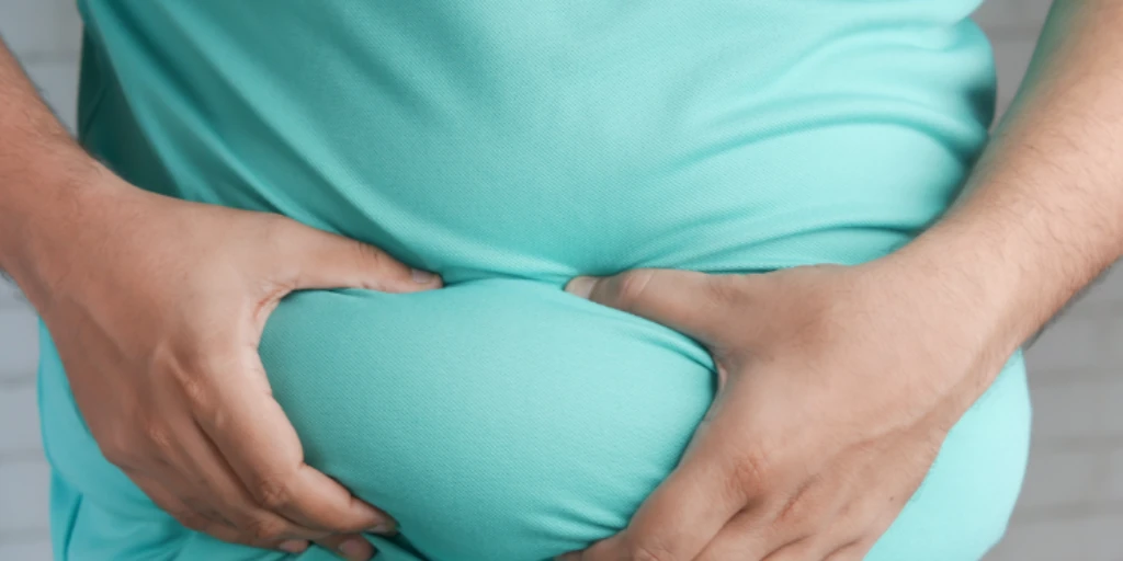 Dicke Person in türkisfarbenen T-Shirt fasst sich mit beiden Händen an sein Speckpolster am Bauch