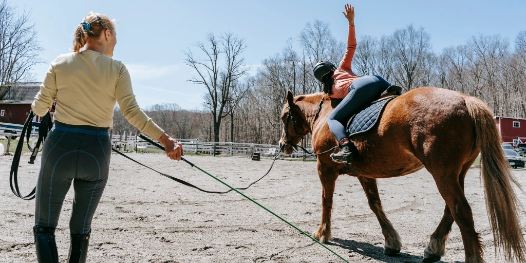 Reitlehrerin mit Peitsche in der rechten Hand und Longe in der Linkenhand hat einen Reitschüler der gerade Gymnastik auf dem Pferd macht.